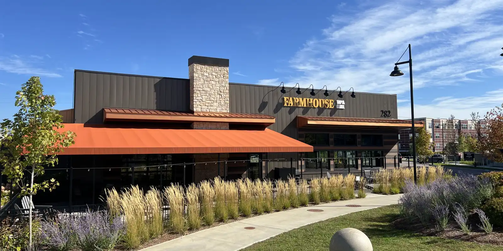 the front of the Farmhouse Williston restaurant with blue sky and flowers in bloom, the outdoor covered patio can be seen